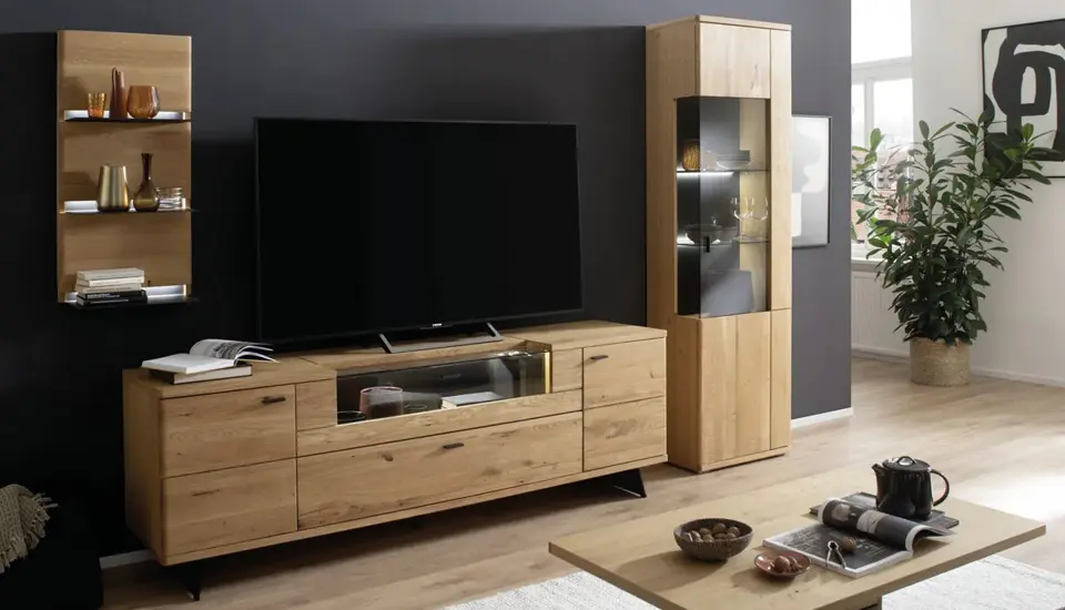 Stilvolle Wohnwand aus massiver Eiche mit großem TV-Board, beleuchteter Vitrine und Wandregal, kombiniert mit modernen Deko-Elementen und dunkler Akzentwand.